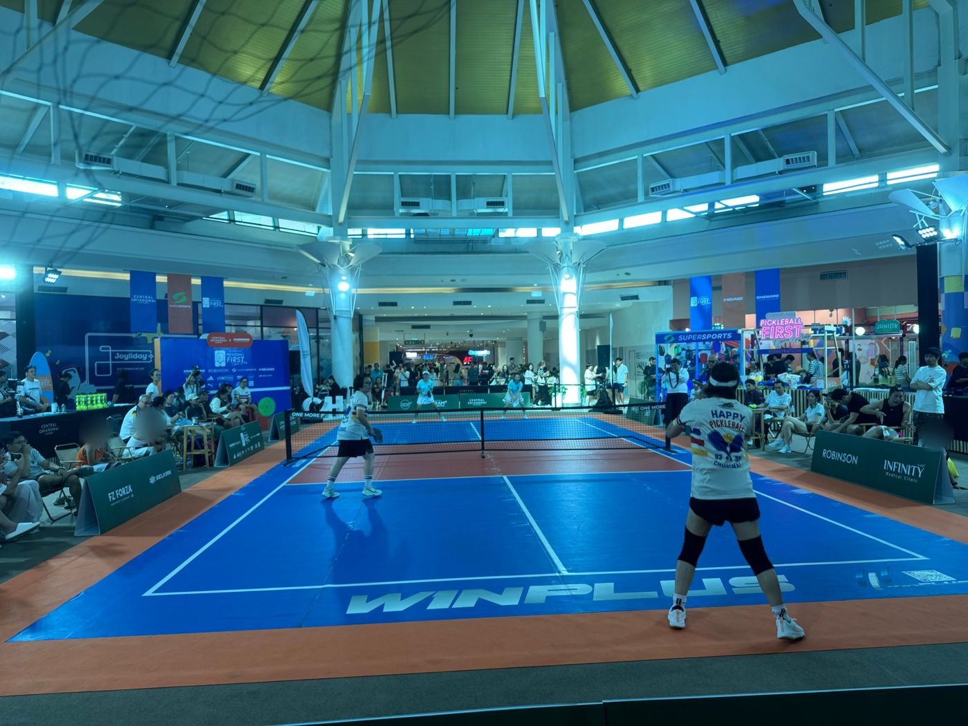 Two players in a doubles match on the blue and orange pickleball court set up under the dome at Central Chiangmai Airport, with spectators seated around the court