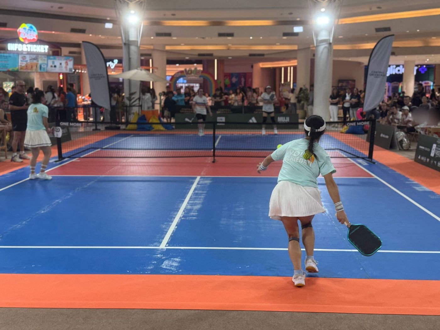 A player stretching for a forehand during a doubles match at Central Chiangmai Airport, with a crowd of shoppers watching beyond the net
